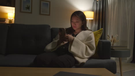 Woman-Spending-Evening-At-Home-Sitting-On-Sofa-With-Mobile-Phone-Scrolling-Through-Internet-Or-Social-Media-6